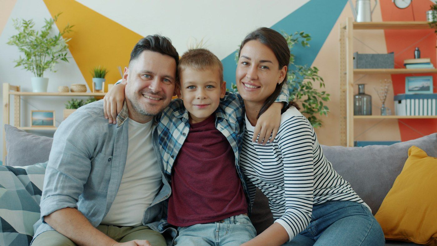 Pai, mãe e filho abraçados e sorrindo no sofá de casa